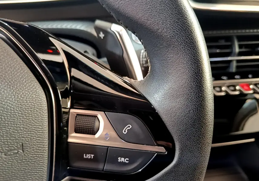 Gros plan sur le volant cuir noir du Peugeot 2008 gris clair, avec commandes audio et tableau de bord en arrière-plan.