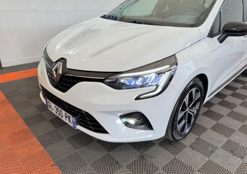 Vue 3/4 avant droite d'une Renault Clio blanche avec phares LED allumés et jantes noires bicolores.