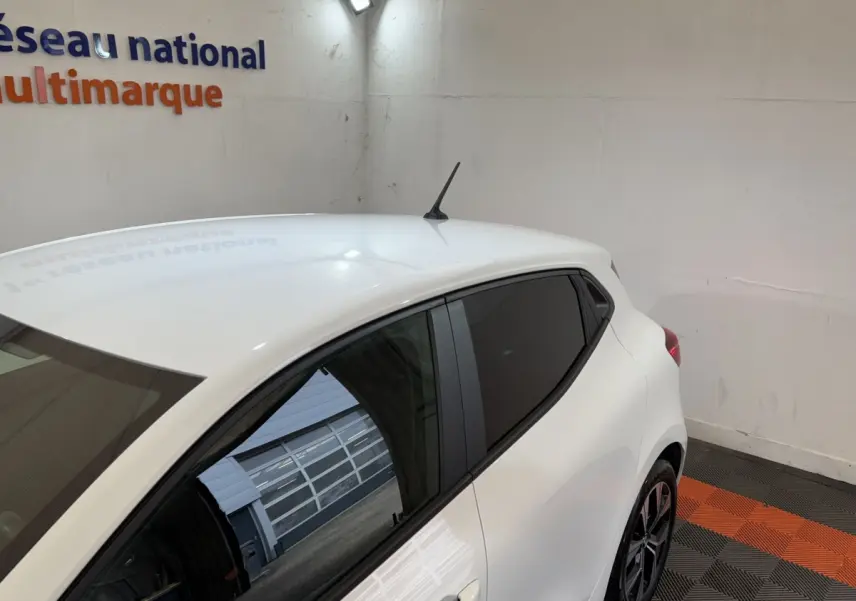 Vue 3/4 arrière droite d'une Renault Clio Business blanche, vitres teintées et jantes noires, dans un garage intérieur.