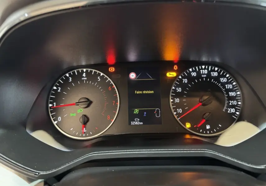 Vue rapprochée du tableau de bord de la Renault Clio blanc 2023, affichant un message "Faire révision" et compteurs analogiques.