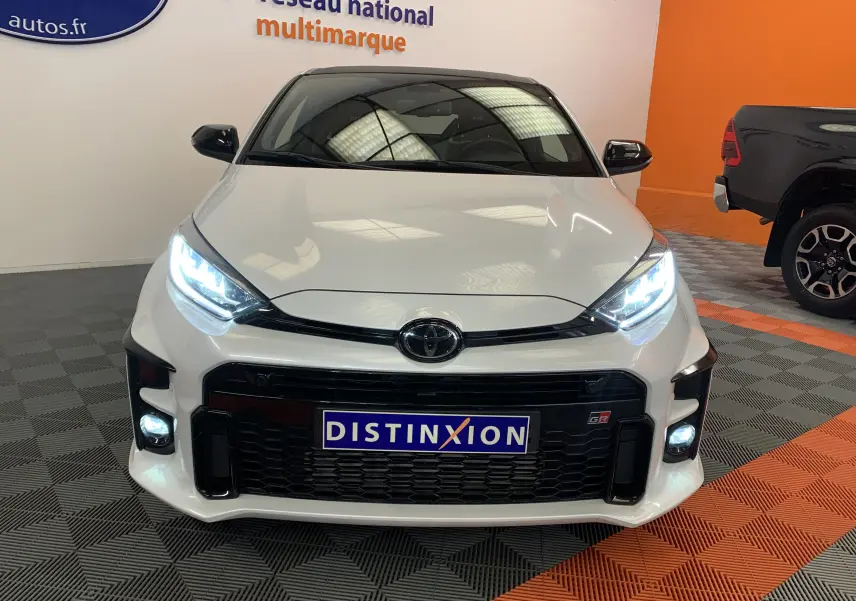 Vue avant d'une Toyota Yaris 1.6 VVT-I 261 Premium GR blanc métal avec phares LED allumés en showroom.