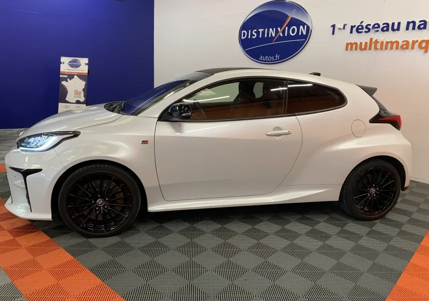 Profil côté gauche d'une Toyota Yaris 1.6 VVT-I 261 Premium GR blanche métal avec jantes noires en showroom.