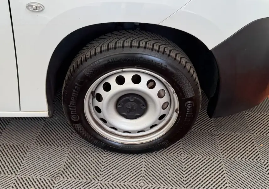 Gros plan sur la roue avant droite acier d’un Peugeot Partner blanc avec pare-chocs noir et sol en damier gris et orange.