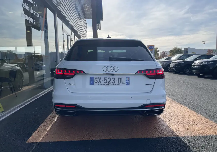 Vue arrière d'une Audi A4 Avant 40 TDI blanche de 2022 avec feux LED allumés, stationnée devant un bâtiment vitré.