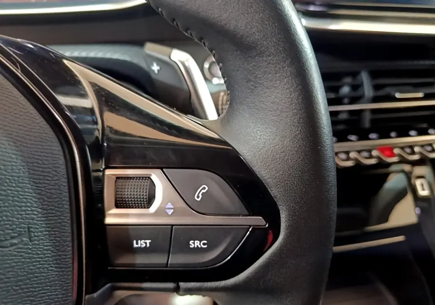 Gros plan sur les commandes gauche du volant cuir noir du Peugeot 2008, avec tableau de bord et écran tactile en arrière-plan.