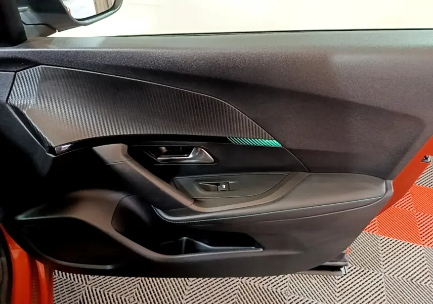 Intérieur côté gauche de la porte avant d’un Peugeot 2008 orange, avec habillage noir et insert décoratif carbone.