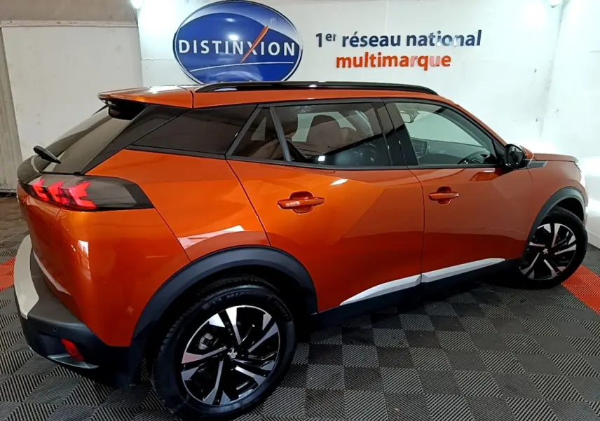 Peugeot 2008 orange vue 3/4 arrière droit, avec toit noir et jantes alliage bicolores, stationné en intérieur.