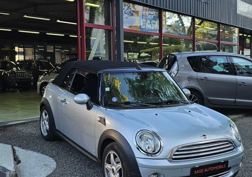 MINI Cabriolet grise vue 3/4 avant droit, capote noire fermée, stationnée devant un garage automobile.