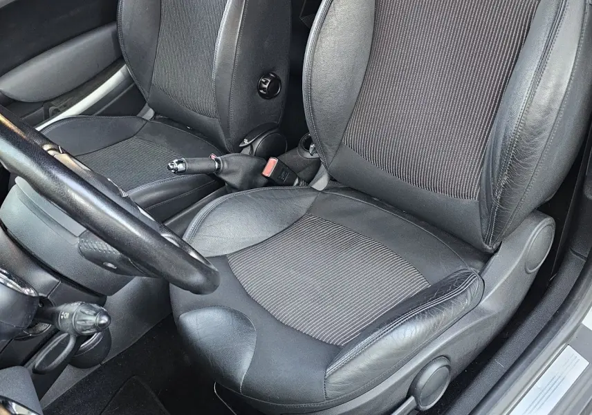 Intérieur côté conducteur d'une MINI Cabriolet R57 grise de 2010, sièges en cuir noir et tissu, volant et frein à main visibles.