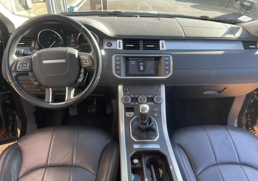 Vue intérieure avant du Range Rover Evoque noir de 2016, montrant le volant, la console centrale avec boîte manuelle et l’écran tactile.