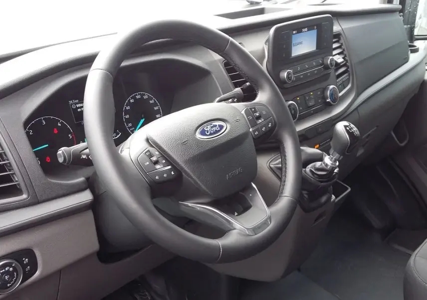 Vue intérieure côté conducteur du Ford Transit Fourgon blanc, avec volant multifonction et levier de vitesses manuel visible.