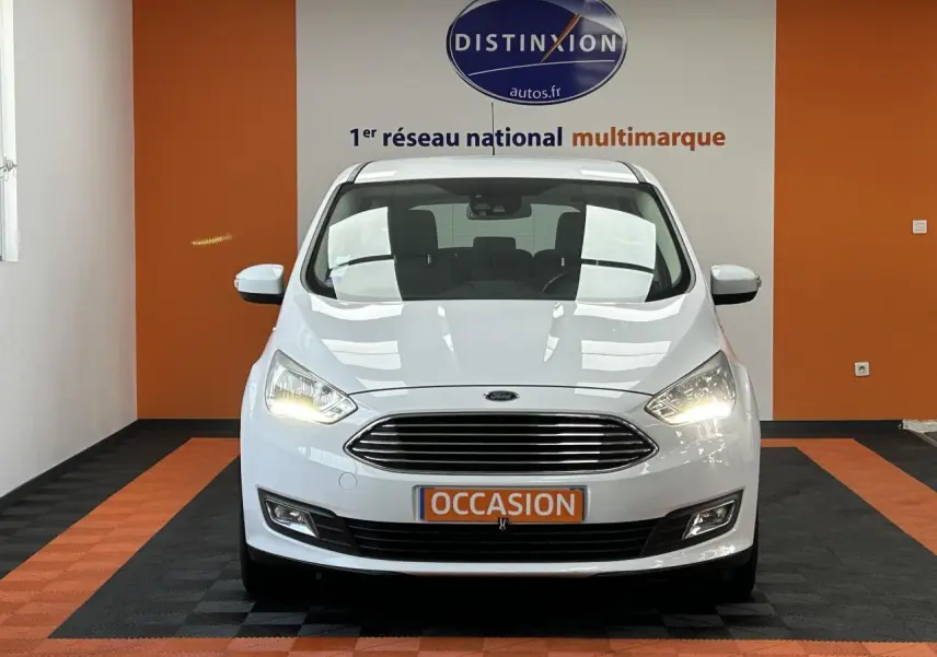 Vue frontale d'une Ford C-MAX blanche 2018 avec calandre chromée et phares allumés dans un showroom.