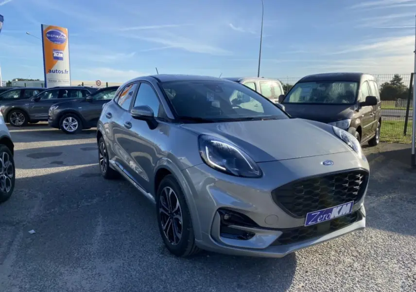 Ford Puma gris en 3/4 avant droit sur parking, avec calandre noire et jantes classiques visibles.