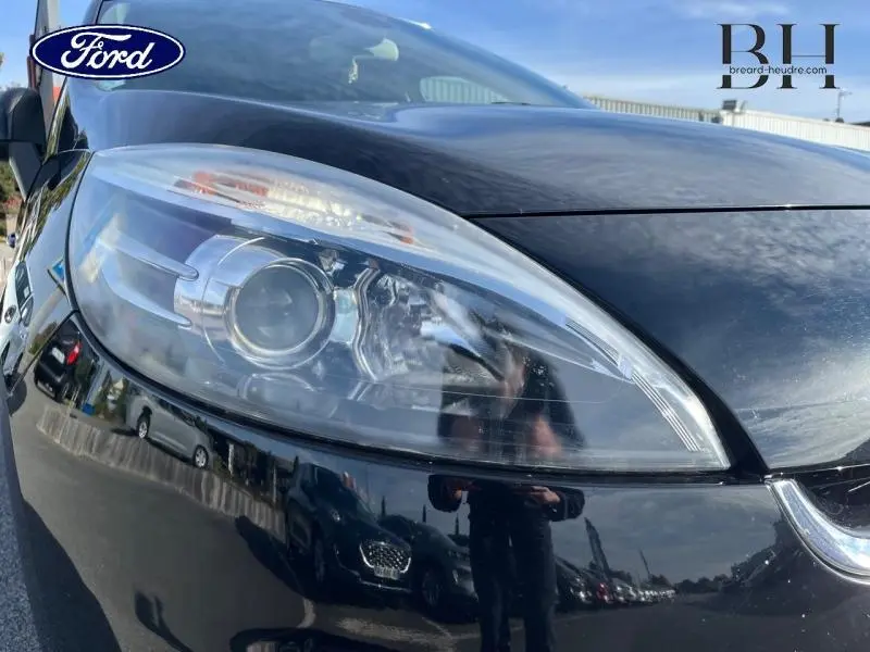 Gros plan sur le phare avant gauche noir étoilé du Renault Grand Scénic 2012 avec reflets du ciel et environnement.