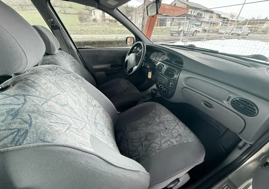 Vue intérieure côté conducteur de la Renault Mégane 2000, montrant les sièges en tissu gris et le tableau de bord simple.