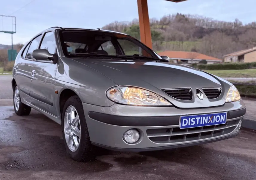 Vue 3/4 avant gauche d'une Renault Mégane grise avec phares allumés et jantes alliage, stationnée sous abri.