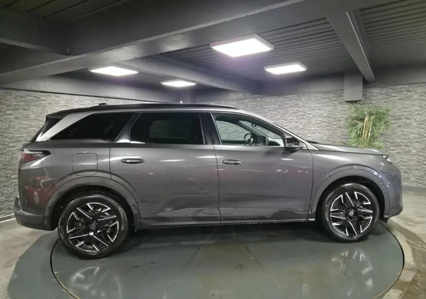 Profil droit d'un Peugeot 5008 gris métallisé 2025 avec jantes alliage noires et toit noir en intérieur showroom.