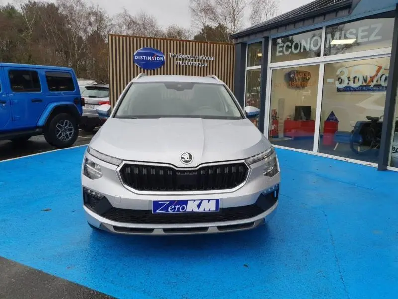 Vue frontale d'un Skoda Kamiq gris diamant métallisé avec calandre noire et phares LED, sur sol bleu devant un showroom.