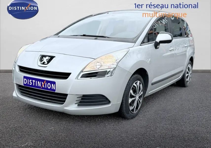 Peugeot 5008 gris vue 3/4 avant droit, montrant la calandre et les phares allumés sur fond neutre.