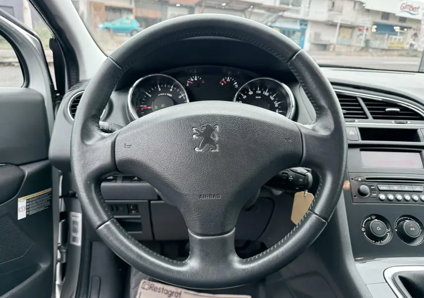 Vue intérieure centrée sur le volant en cuir noir avec logo Peugeot, tableau de bord et commandes de climatisation visibles.