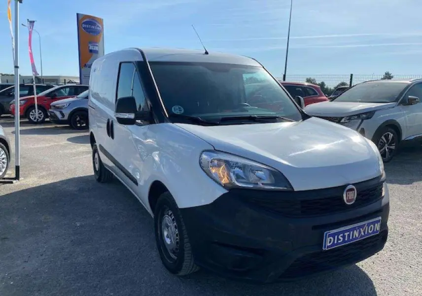 Fiat Doblo blanc vu en 3/4 avant droit, fourgon tôlé avec pare-chocs noirs et porte latérale droite visible.