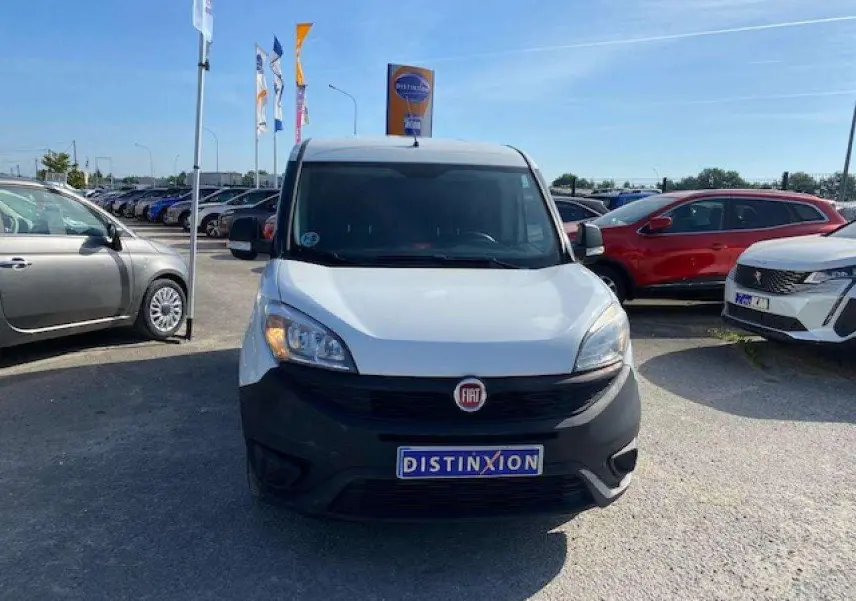Vue avant d'un FIAT Doblo blanc 2017 avec pare-chocs noir et plaque Distinxion sur un parking.