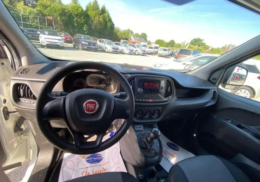 Vue intérieure du poste de conduite du FIAT Doblo blanc de 2017, avec volant, tableau de bord et levier de vitesses manuelle visibles.