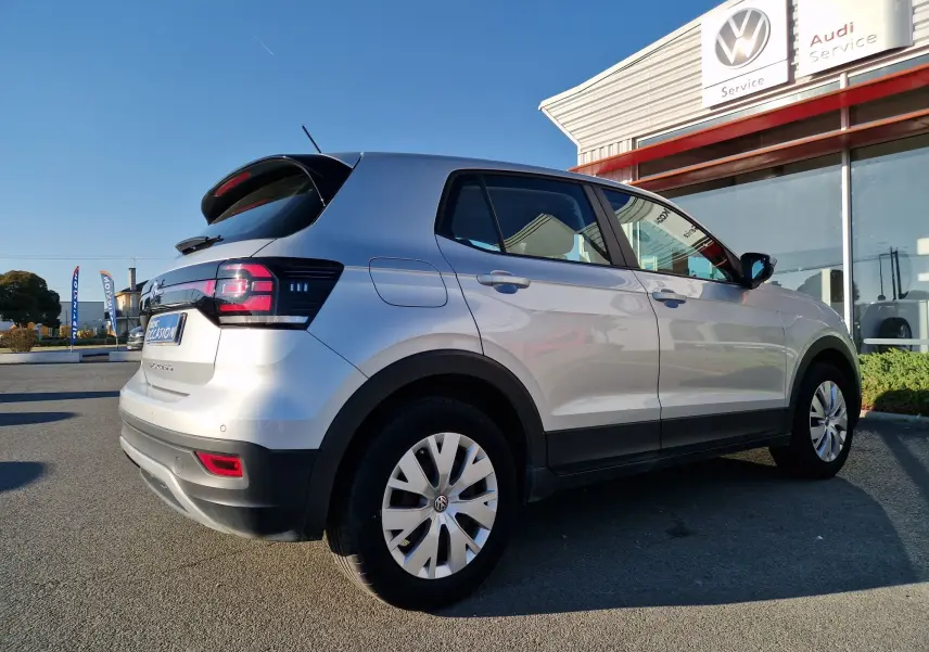Volkswagen T-Cross argent métal vu en 3/4 arrière droit, stationné devant un service Volkswagen et Audi.
