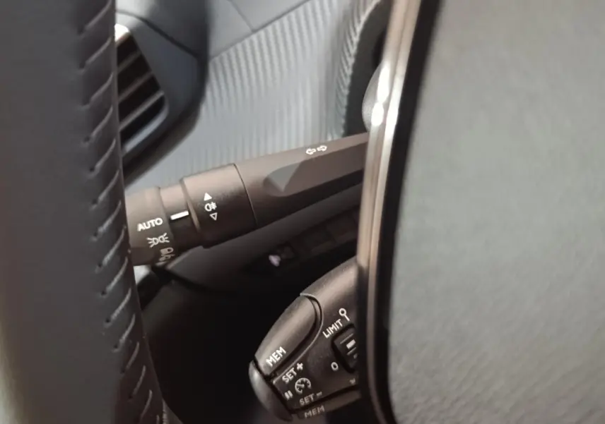 Gros plan sur les commandes au volant cuir de la Peugeot 208 électrique 2024, avec réglages du régulateur et feux automatiques.