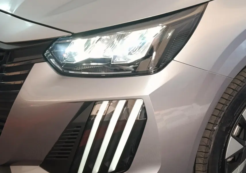 Gros plan sur l'avant droit gris clair du Peugeot 208 électrique 2024, mettant en valeur ses phares LED et feux diurnes verticaux.