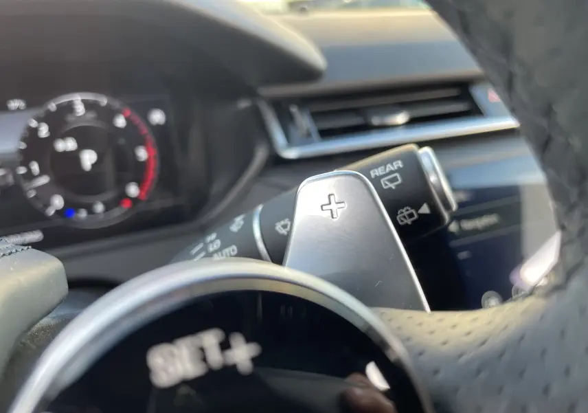 Gros plan sur la palette de changement de vitesse + au volant perforé du Range Rover Velar noir Santorini, tableau de bord flou en arrière-plan.