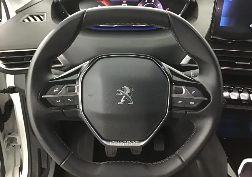 Vue rapprochée du volant cuir noir du Peugeot 5008 blanc nacré, avec commandes intégrées et tableau de bord numérique.