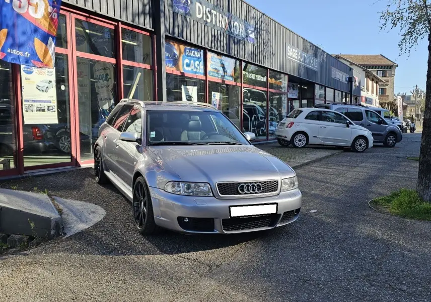 Audi RS4 Avant gris vue 3/4 avant droit, avec jantes noires et calandre distinctive, stationnée devant un atelier.