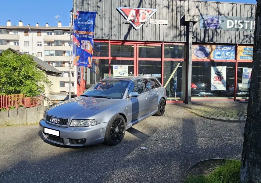 Audi RS4 Avant gris en 3/4 avant droit, avec jantes noires, stationnée devant un garage ensoleillé.