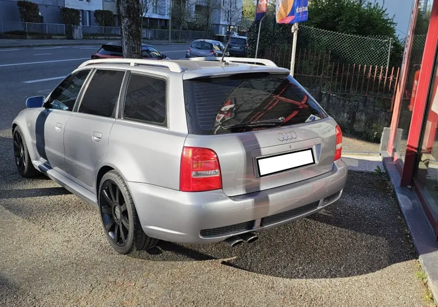Audi RS4 Avant gris vue 3/4 arrière droit, avec jantes noires et toit ouvrant, stationnée près d'un bâtiment.