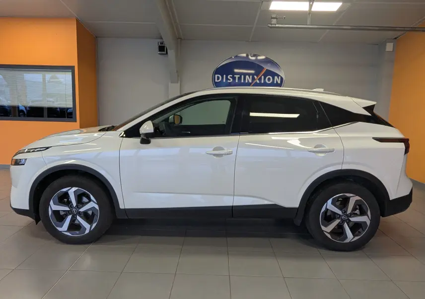 Vue latérale droite du Nissan Qashqai 2024 blanc, avec jantes bi-ton et toit noir contrastant en intérieur showroom.