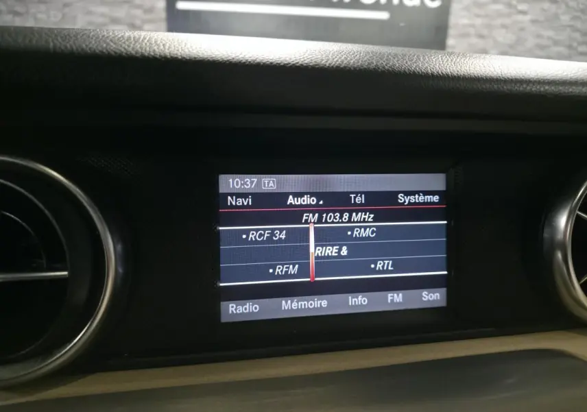 Écran central de navigation et audio affichant la fréquence FM 103.8 MHz dans l'habitacle d'une Mercedes SLK 250 CDI gris