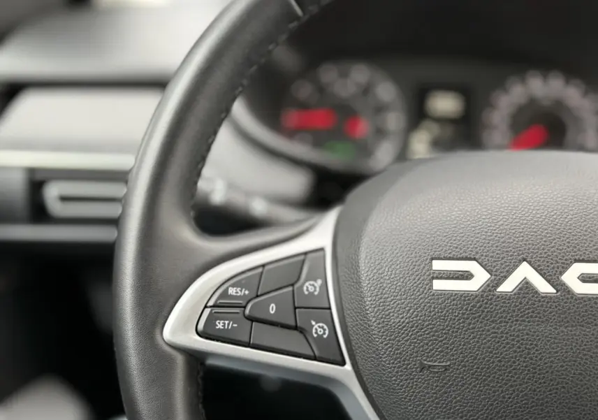 Gros plan sur le volant noir de la Dacia Jogger 2025 avec commandes de régulateur de vitesse, tableau de bord flou en arrière-plan.