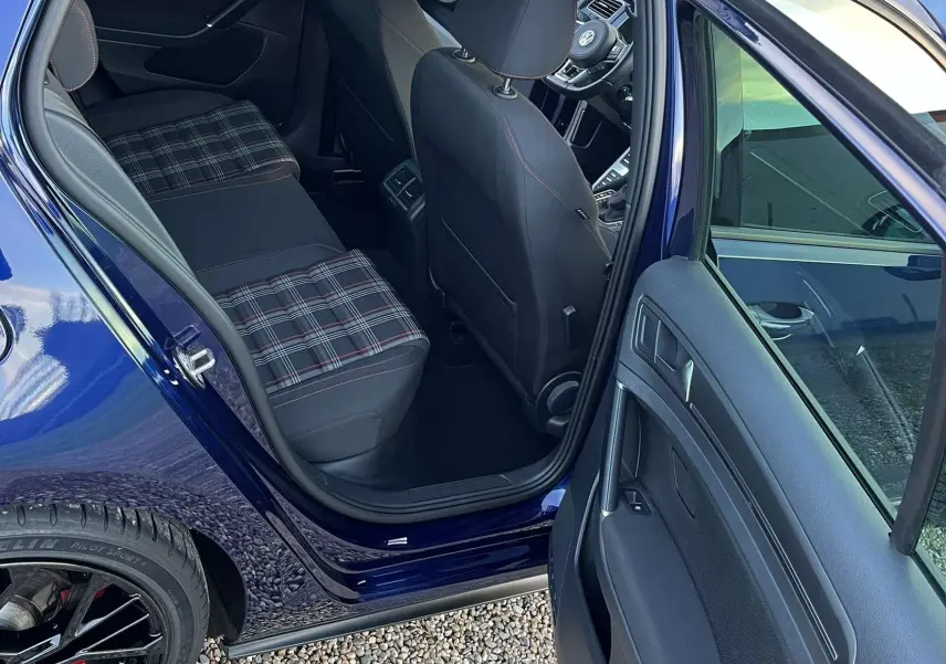 Vue intérieure côté arrière gauche d'une Volkswagen Golf VII GTI Performance bleu lapiz, sièges à motif tartan et volant visible à l'avant.