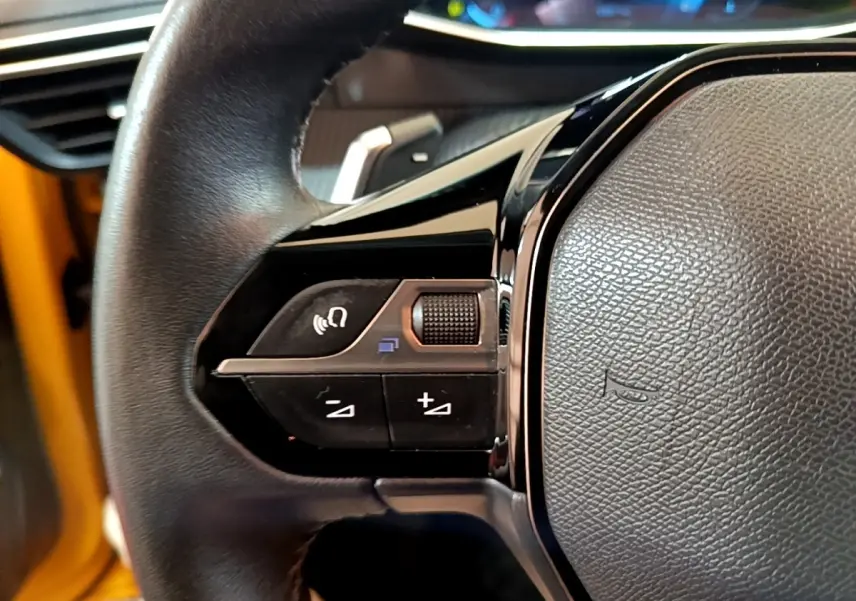 Gros plan sur le volant cuir de la Peugeot 208 jaune, avec commandes multifonctions et tableau de bord digital en arrière-plan.