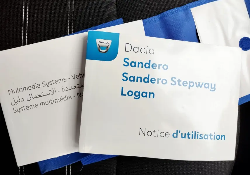 Notice d'utilisation Dacia Sandero Stepway Logan posée sur un siège noir avec surpiqûres blanches et un tissu bleu visible.
