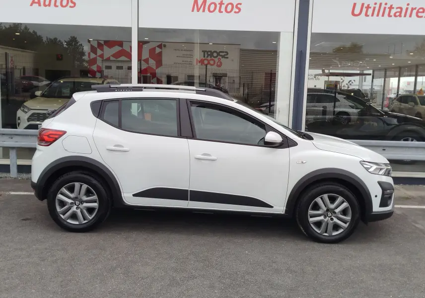 Vue latérale droite d'une Dacia Sandero Stepway blanche Glacier avec barres de toit et protections noires sur les bas de caisse.