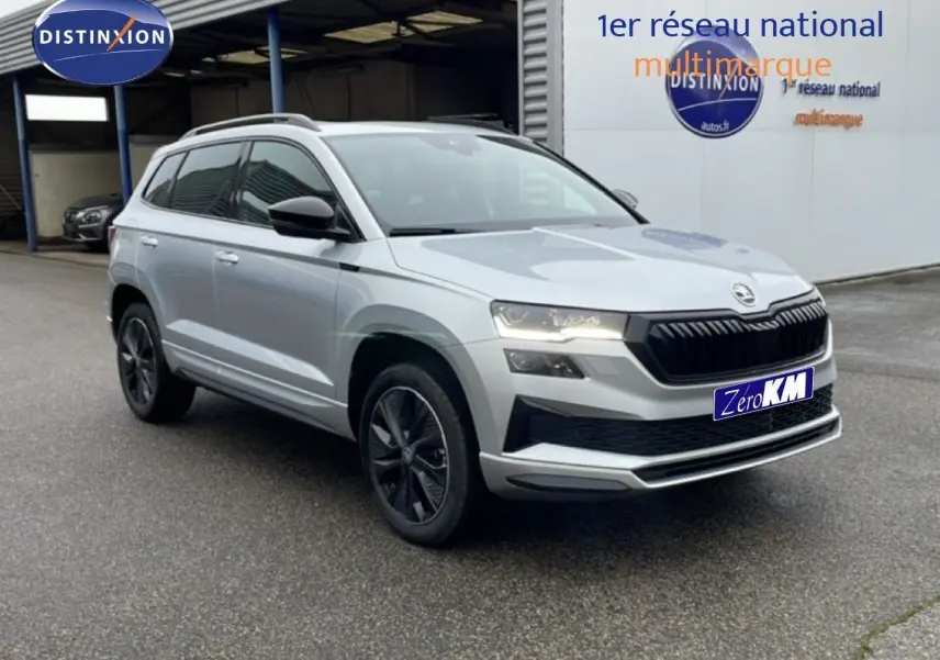 SUV Skoda Karoq gris argent métal en 3/4 avant droit, avec calandre noire et jantes foncées, stationné devant un garage.