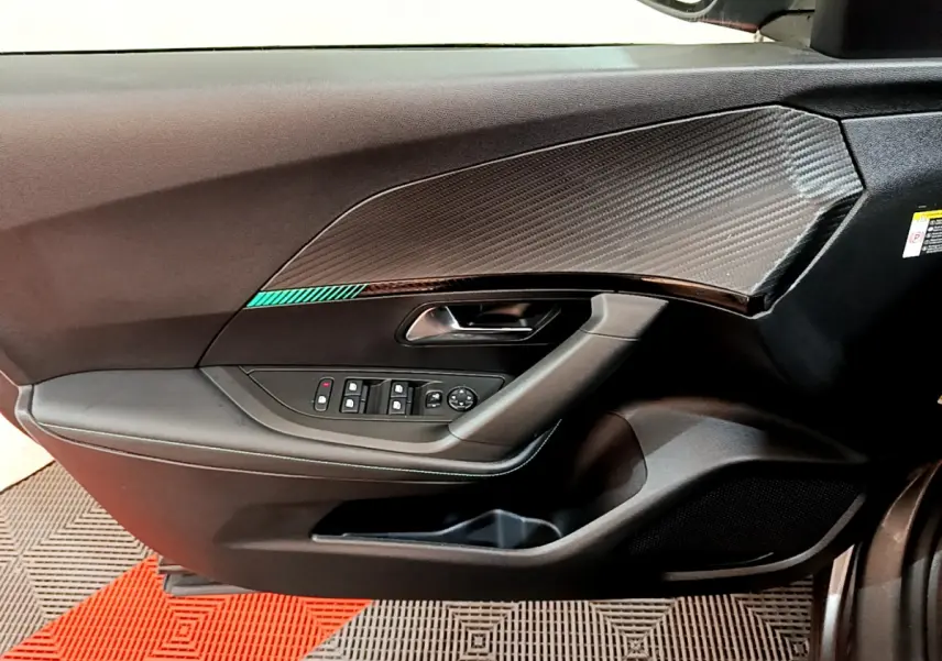 Vue intérieure côté gauche de la porte d'une Peugeot 2008 gris foncé, avec garniture carbone et commandes électriques.