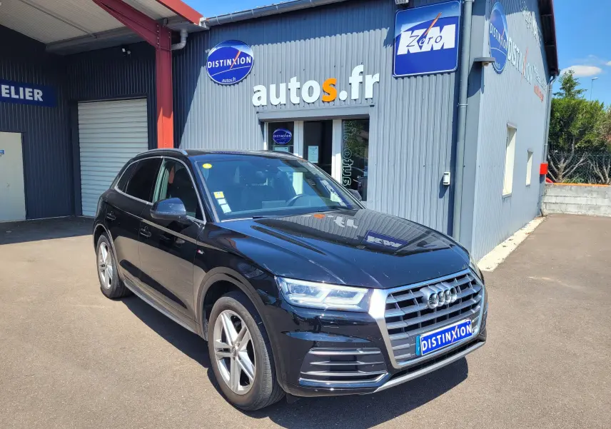 Audi Q5 noire vue en 3/4 avant droit, stationnée devant un garage avec logo autos.fr visible en arrière-plan.