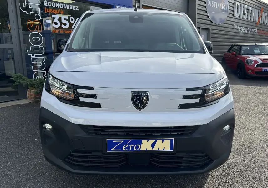 Vue de face d'un Peugeot Partner Fourgon blanc avec calandre noire et plaque Zéro KM, stationné devant un garage.