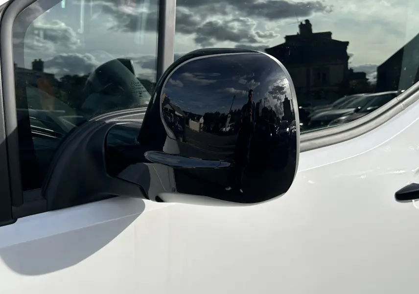 Rétroviseur noir côté conducteur du Peugeot Partner Fourgon blanc 2025, avec reflet urbain sur fond de ciel nuageux.