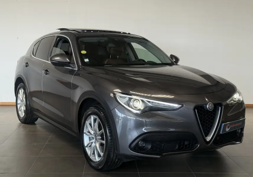 Alfa Romeo Stelvio gris en 3/4 avant droit, avec calandre caractéristique en V et phares allumés.