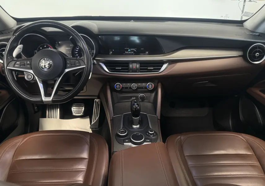Vue intérieure de l'Alfa Romeo Stelvio 2017 avec tableau de bord noir, sièges cuir marron et console centrale bois.