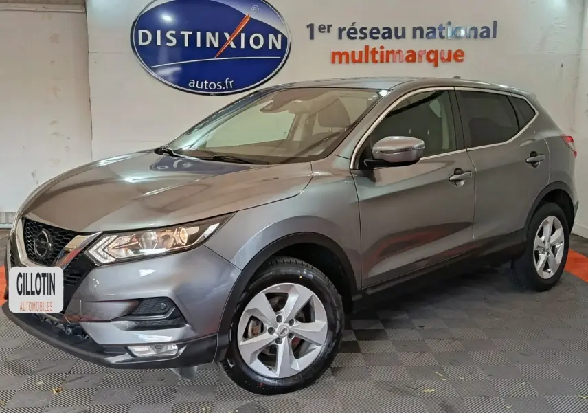 Nissan Qashqai gris clair vu de 3/4 avant droit, avec jantes alliage et phares allumés en intérieur showroom.